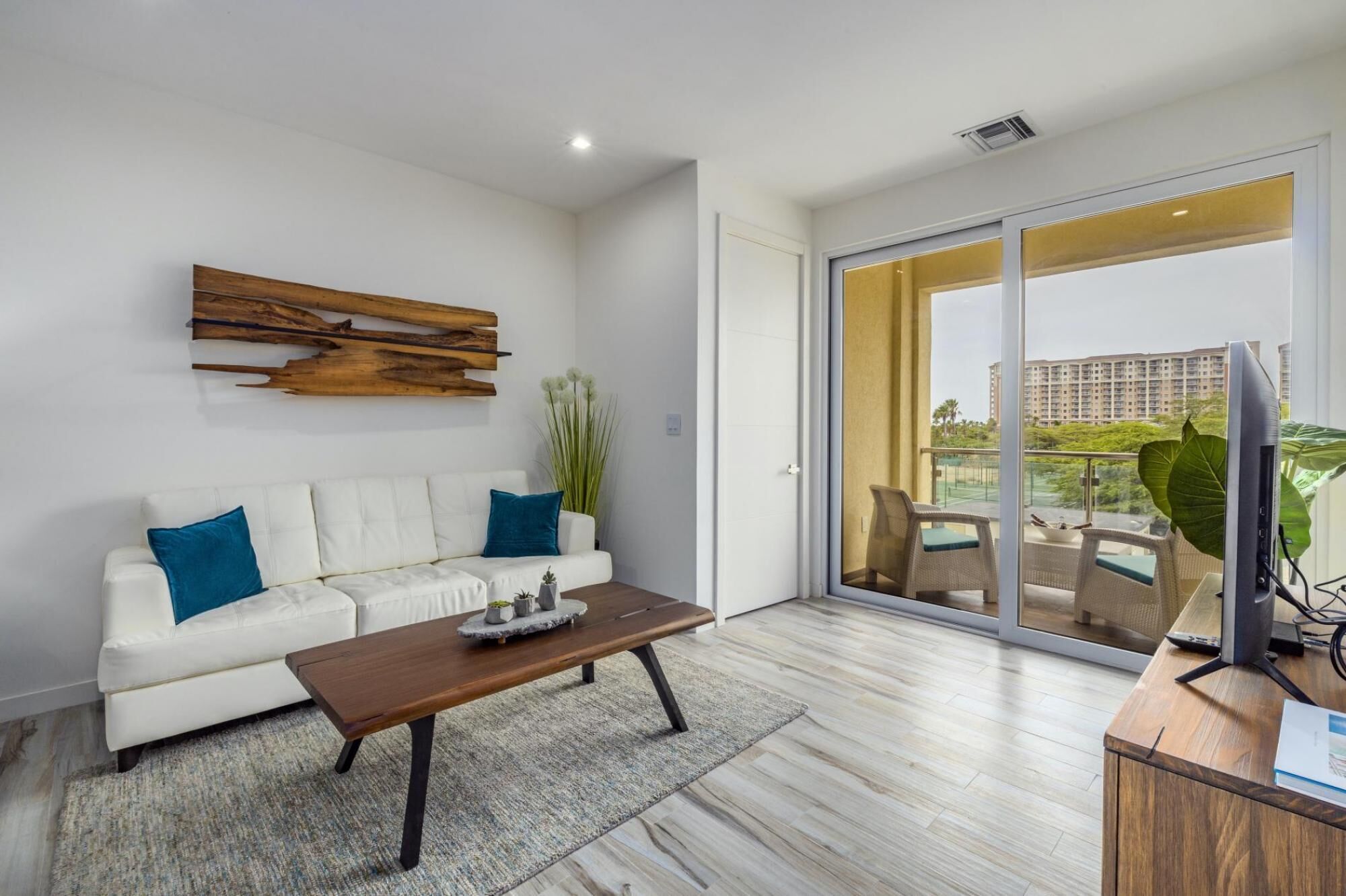Una sala de estar moderna con un sofá blanco, una mesa de centro de madera y un gran ventanal corredero que da a un balcón con vistas a un edificio de gran altura.