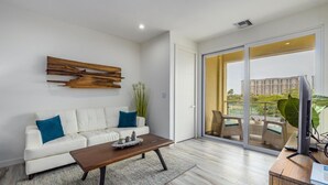 A modern living room with a white sofa, a wooden coffee table, and a large sliding glass door leading to a balcony with a view of a high-rise building. - NEW 2BR Condo in of Palm Beach Walk 2 Beach (Noord)