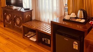Habitación de hotel con piso de madera, un televisor en un mueble bajo, un refrigerador pequeño, un banco y una mesa con una tetera y tazas.