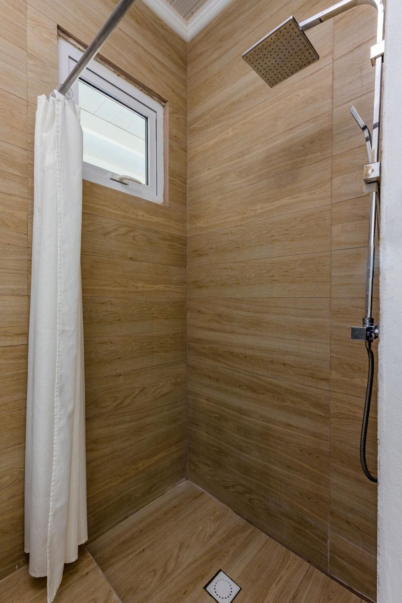 Un espace douche avec des murs en bois, un rideau de douche blanc, une douchette à main et une fenêtre.