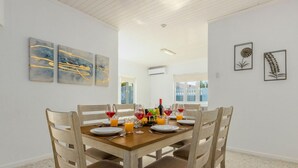 A dining area with a wooden table set for six, surrounded by chairs. There are artworks on the walls, a window with a view, and an air conditioning unit on the ceiling. - 3BR Home w Pool in Downtown 5min Walk 2 Beach (Oranjestad)