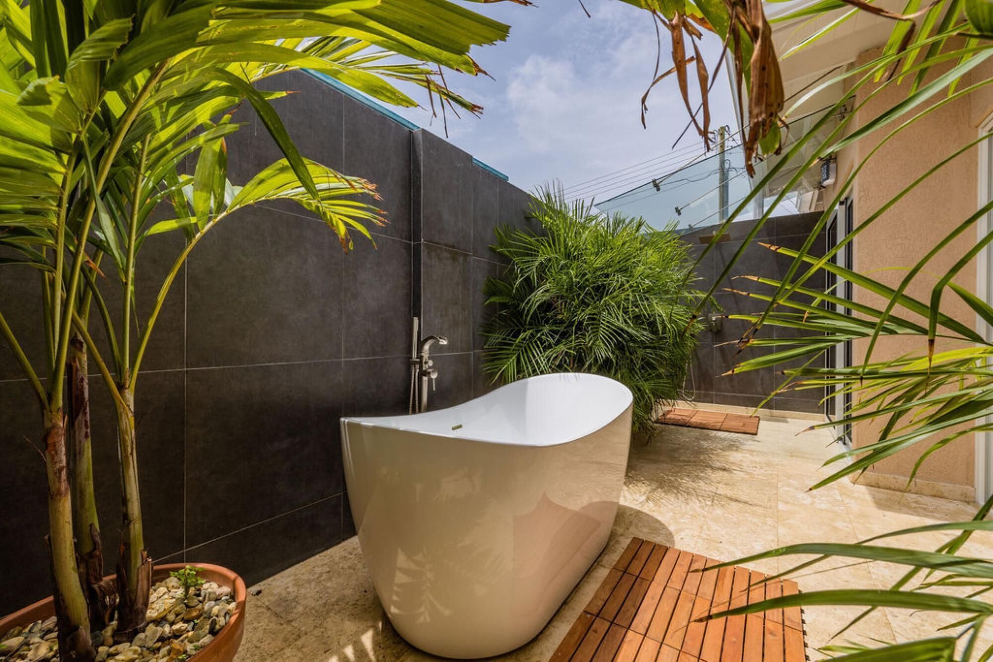 A modern outdoor bathroom with a freestanding bathtub, surrounded by tropical plants and a dark wall.