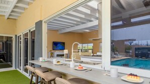 Una cocina moderna con un amplio ventanal con vistas a una piscina, taburetes de bar y un comedor con chimenea.