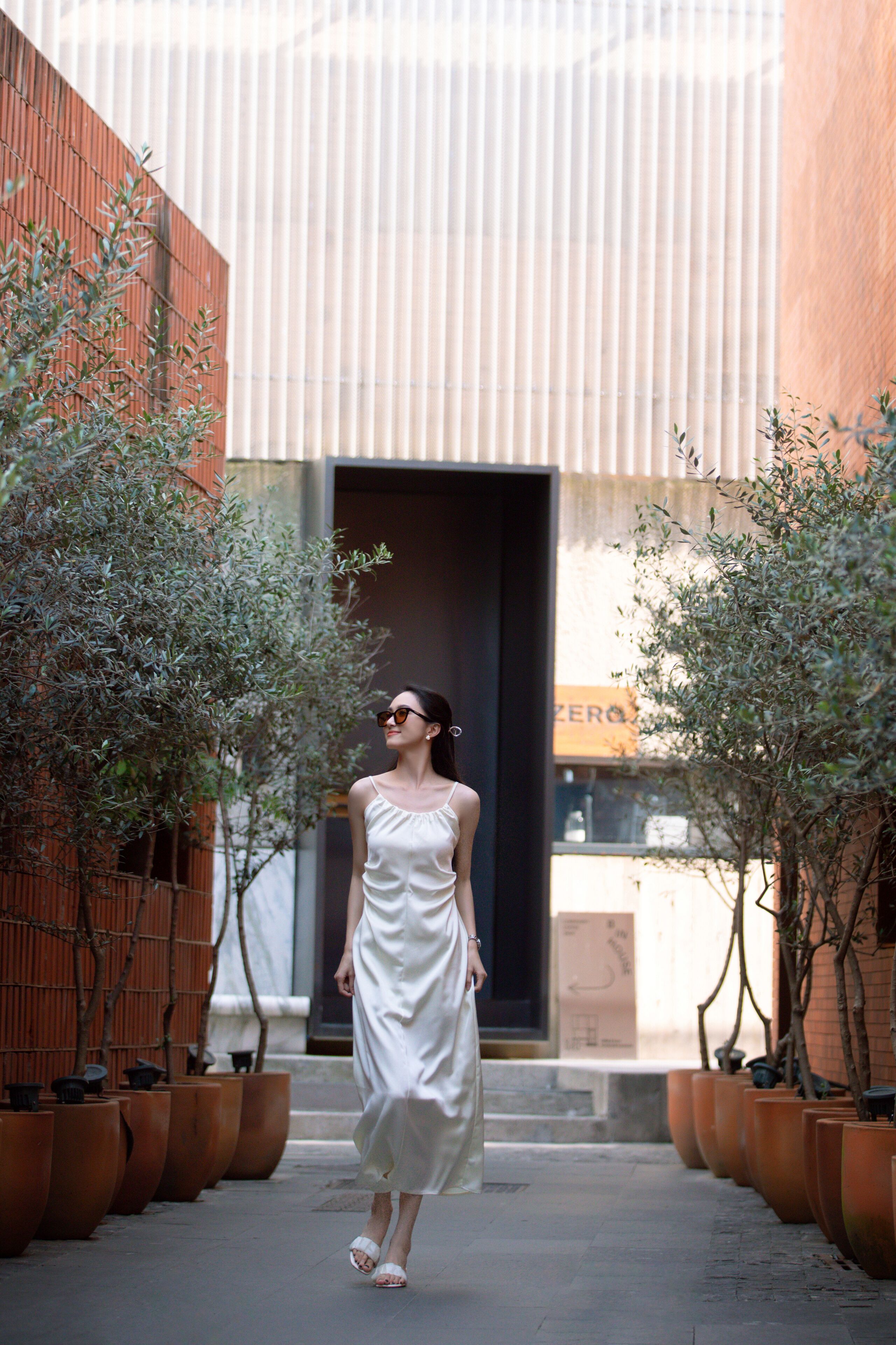 A woman in a white dress walking on a sidewalk with potted plants on either side.
