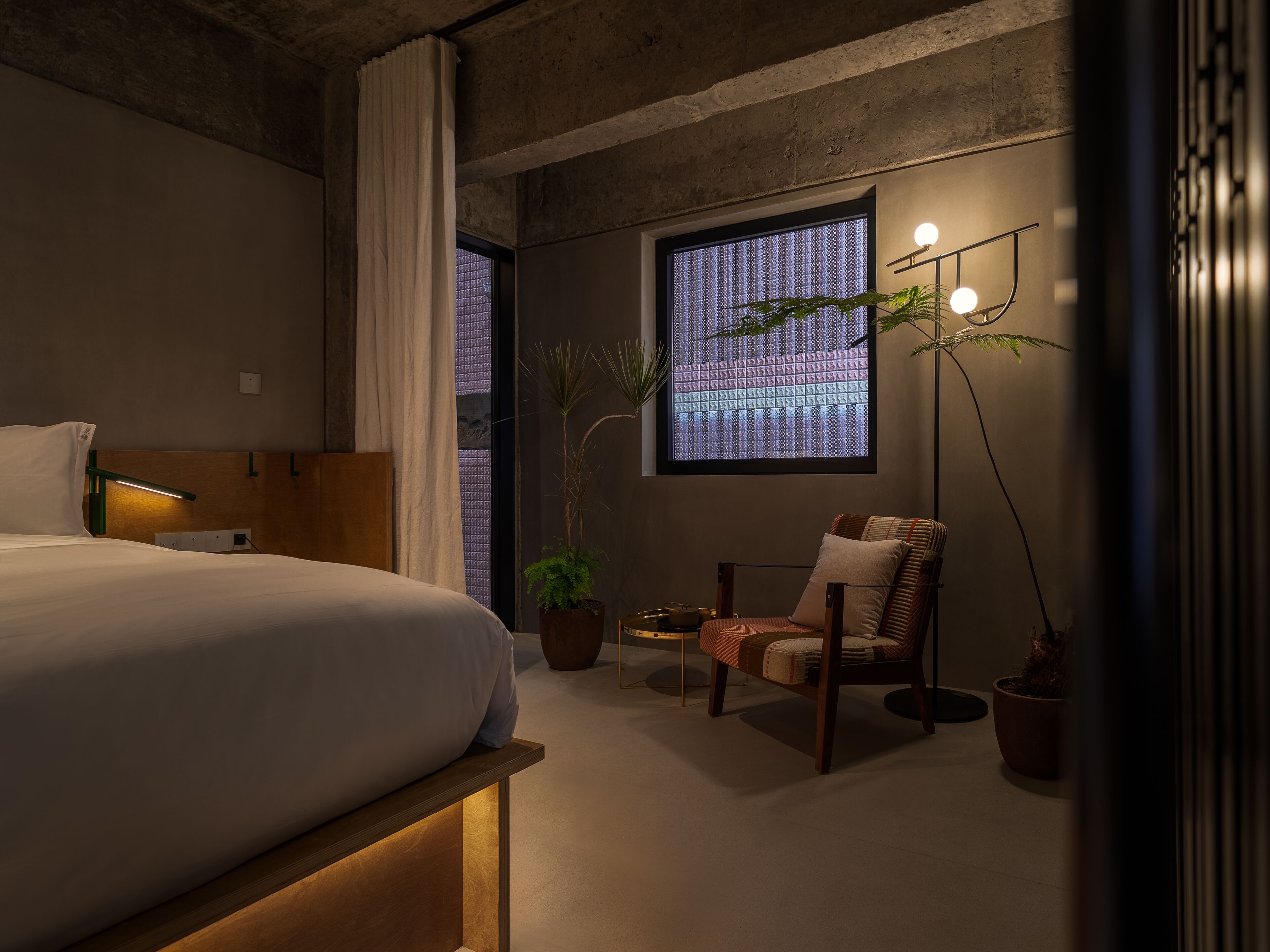 A modern bedroom with a bed, a chair, a floor lamp, and a window with a view of a building.