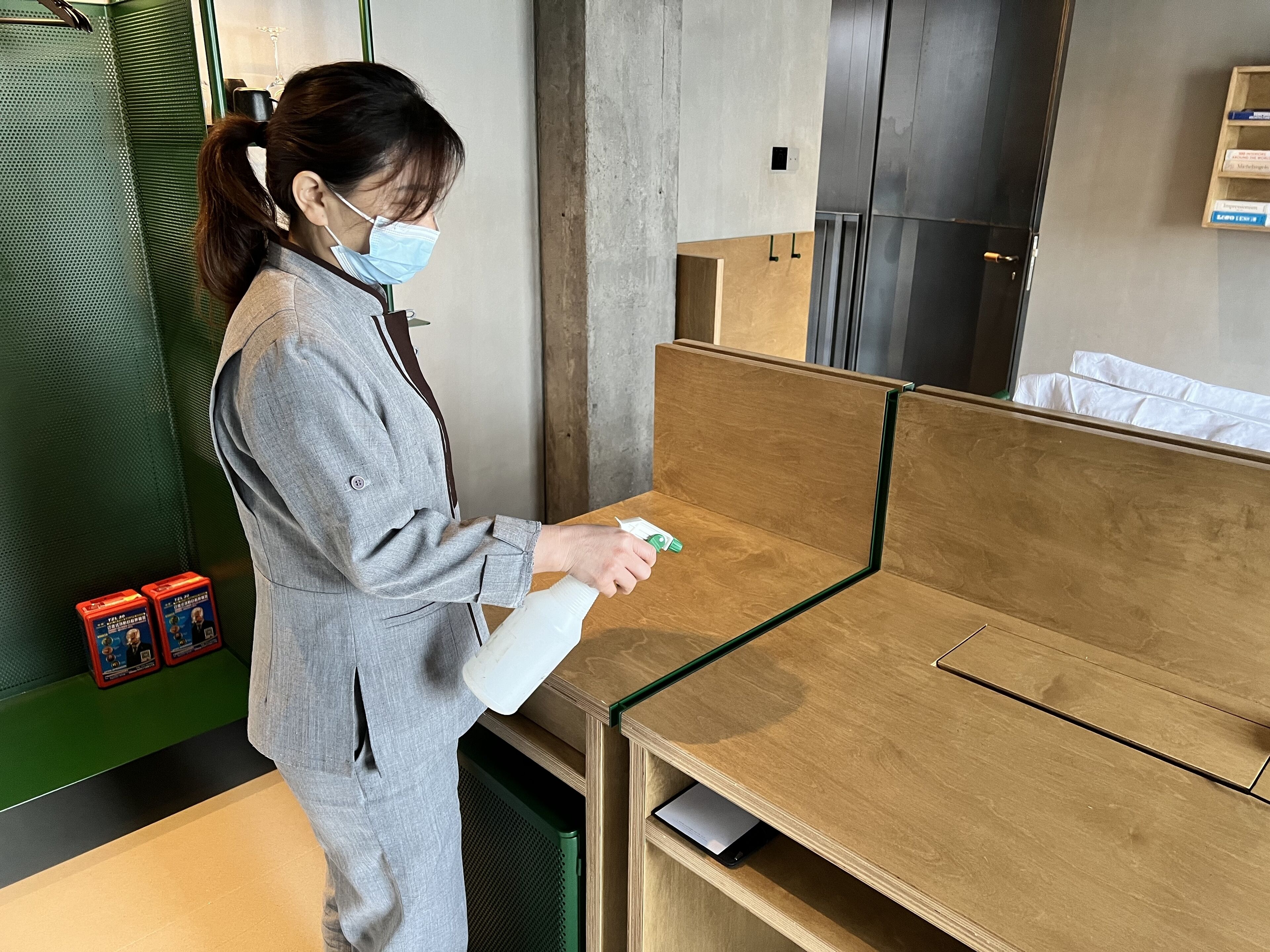 Eine Person in grauer Uniform sprüht eine Fläche in einem Raum mit Holzmöbeln und grüner Wand.