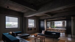 Un salon moderne avec un canapé d’angle, une table basse ronde et un coin repas comprenant une table et des chaises. De grandes fenêtres laissent entrer la lumière naturelle et offrent une vue sur la ville.