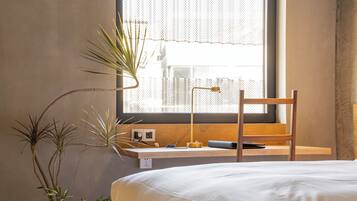 Ein Schlafzimmer mit Bett, Schreibtisch mit Lampe und einem Fenster mit Blick auf Gebäude.