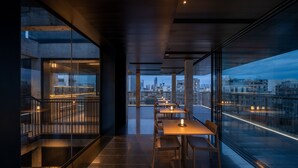 Un comedor moderno con mesas y sillas, ventanas amplias con vistas a la ciudad, y un balcón con barandilla.