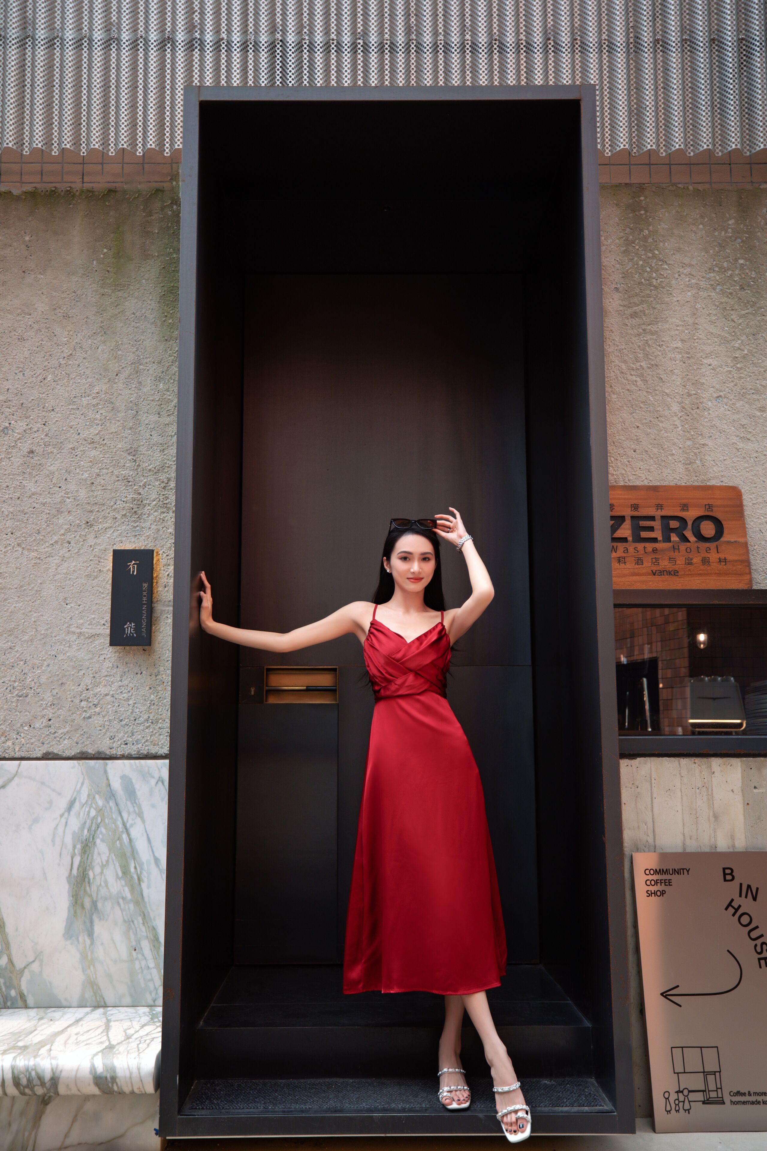 A woman in a red dress standing in a doorway with a sign reading "ZERO Waste Hotel" and a "Community Coffee Shop" sign nearby.