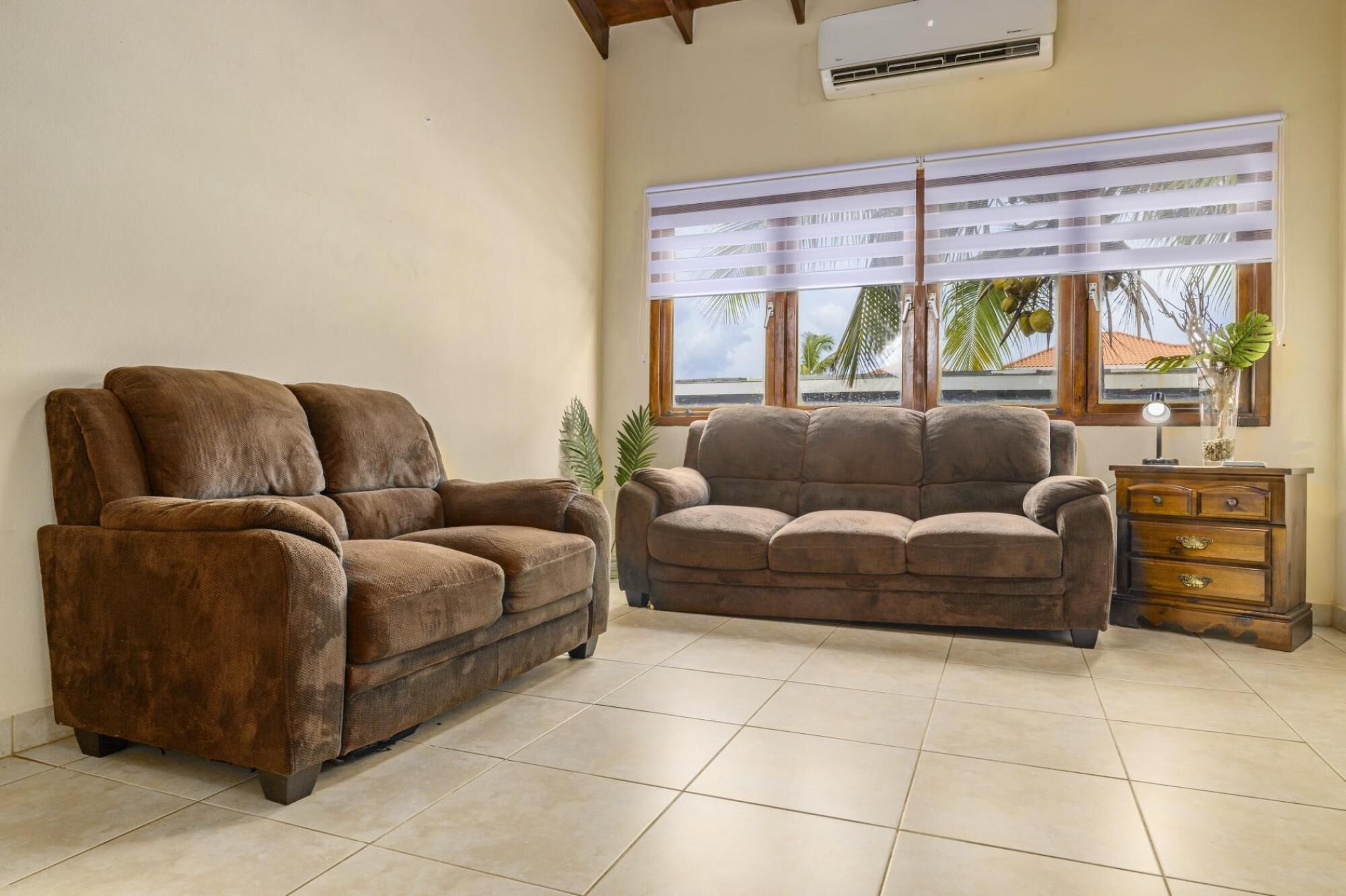 A living room with two brown sofas, a wooden cabinet, and a large window with a view of palm trees.