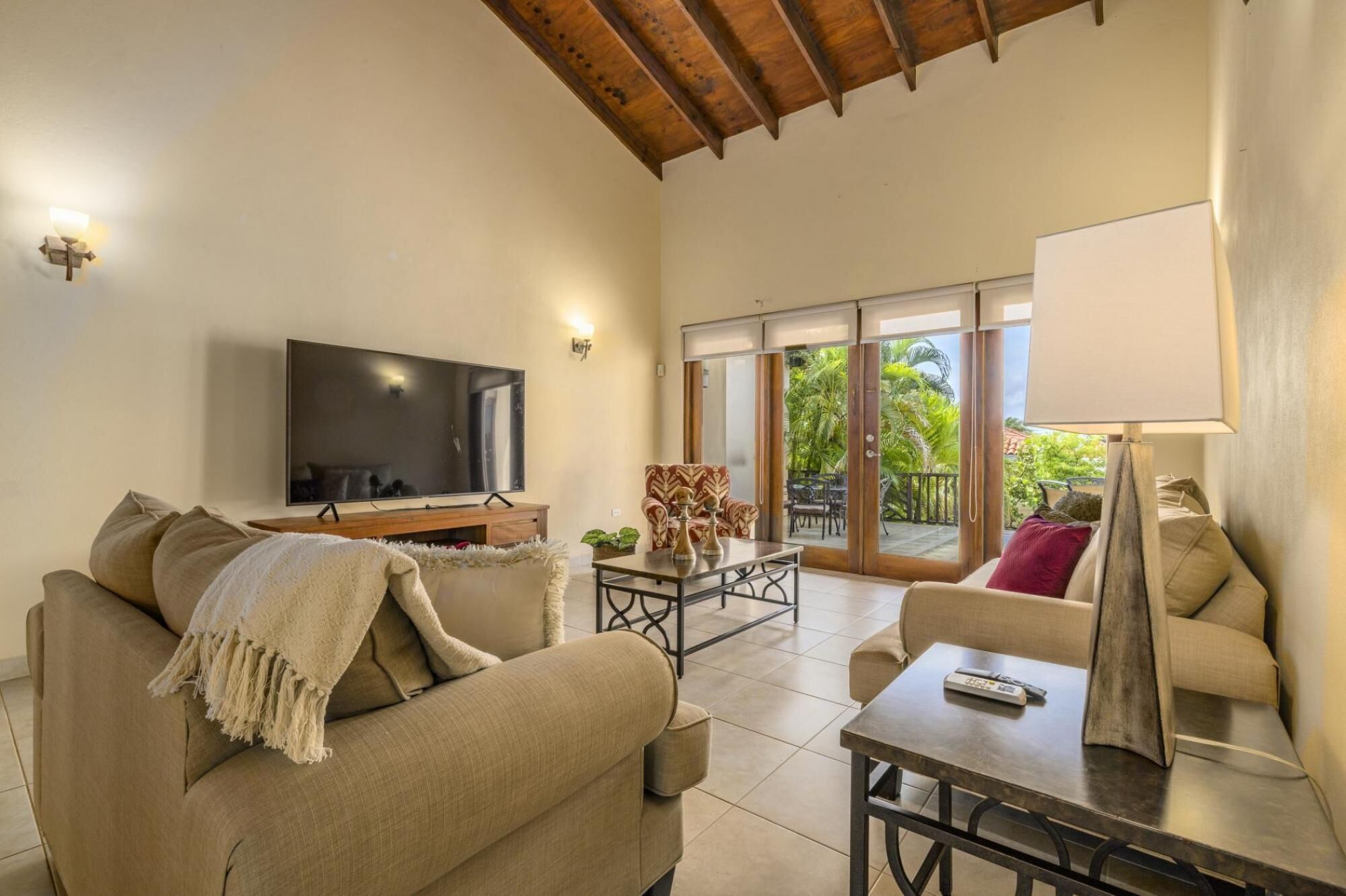 A spacious living area with a sofa, armchair, and a coffee table. A flat-screen TV is mounted on the wall, and there is a sliding glass door leading to an outdoor patio.