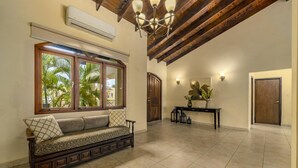 Amplia sala de estar con un banco de madera, un ventanal con vista a palmeras, un ventilador de techo y un aire acondicionado instalado en la pared.