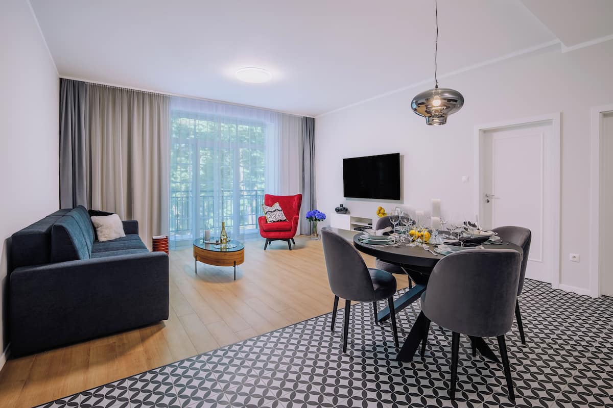 A modern living room with a sofa, dining table, and chairs. There is a TV mounted on the wall and a window with curtains.