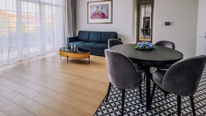 A modern living room with a sofa, a dining table with chairs, a coffee table, and a large window with sheer curtains. - Rezidencia OPERA (Turcianske Teplice)