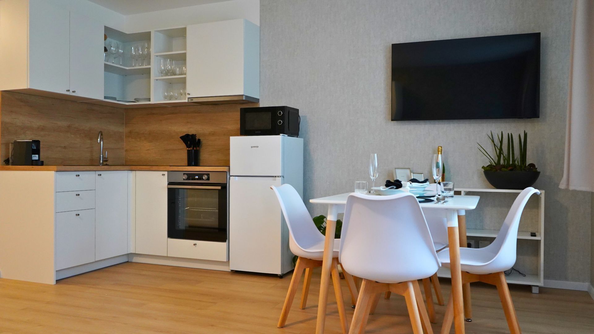 Una cocina moderna con armarios blancos, microondas, horno y refrigerador. Hay una zona de comedor con sillas blancas y una mesa dispuesta para dos.