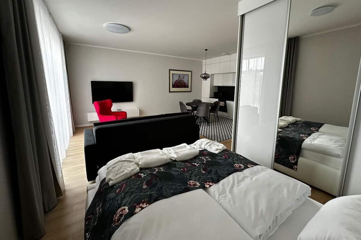 A modern hotel room with a large bed, a desk, a red chair, and a kitchen area.