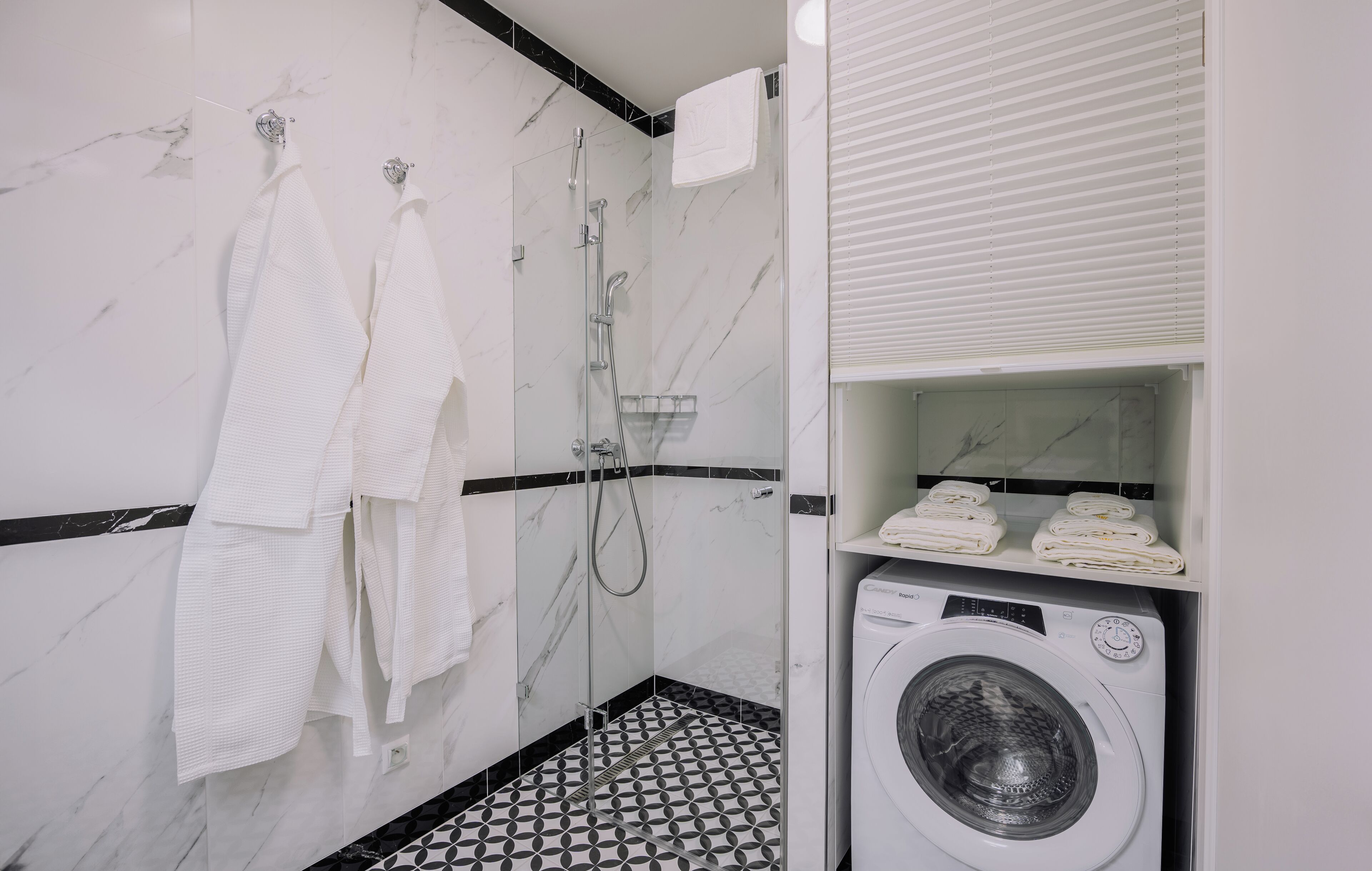 Un baño con pared de mármol, suelo de baldosas blanco y negro, ducha con cerramiento de vidrio y lavadora.