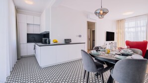A modern kitchen with white cabinets, a black countertop, and a patterned floor. There is a dining area with a table set for four, a red chair, and a grey chair. - Rezidencia OPERA (Turcianske Teplice)