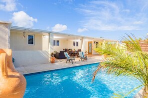 A poolside area with a clear blue pool, sun loungers, a dining table, and a small garden area with a palm tree. - 3BR Privatepool - Outdoor Dining - Great Location (Oranjestad)
