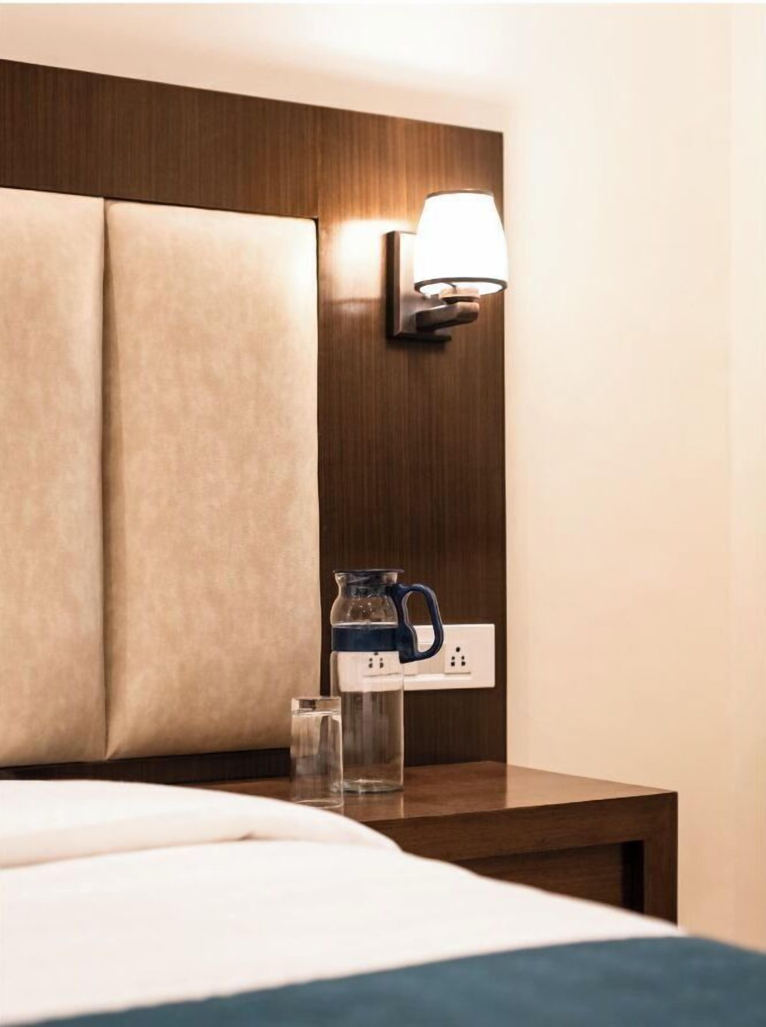 Une chambre d’hôtel avec un lit, une table de chevet, une lampe fixée au mur, une carafe et un verre d’eau.