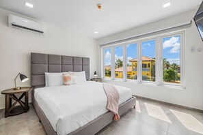Um quarto com uma cama grande, cabeceira cinza, mesa de cabeceira com abajur e janela com vista para uma casa amarela e palmeiras.