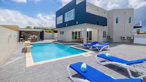 A modern pool area with a rectangular pool, lounge chairs, and a building with large windows. - NEW 6 Bedroom Villa 2min Eagle Beach Palm Beach (Noord)