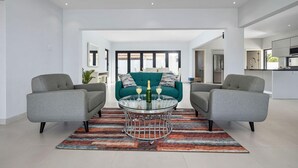 A modern living room with two grey armchairs, a teal sofa, and a glass coffee table with two champagne bottles.