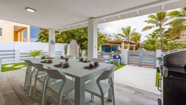 An outdoor patio with a dining table and chairs, a grill, and a view of a pool and palm trees. - Brand NEW 3BR 2BA Villa With Amazing Amenities (Noord)