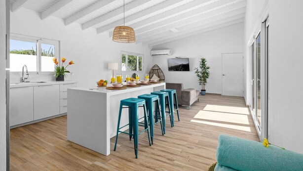 Une cuisine moderne avec des meubles blancs, un îlot central doté de tabourets, un luminaire suspendu et une grande fenêtre laissant entrer la lumière naturelle.