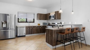Een moderne keuken met een roestvrijstalen koelkast, een magnetron en een fornuis. Er zijn houten kastjes en een raam met zonwering.