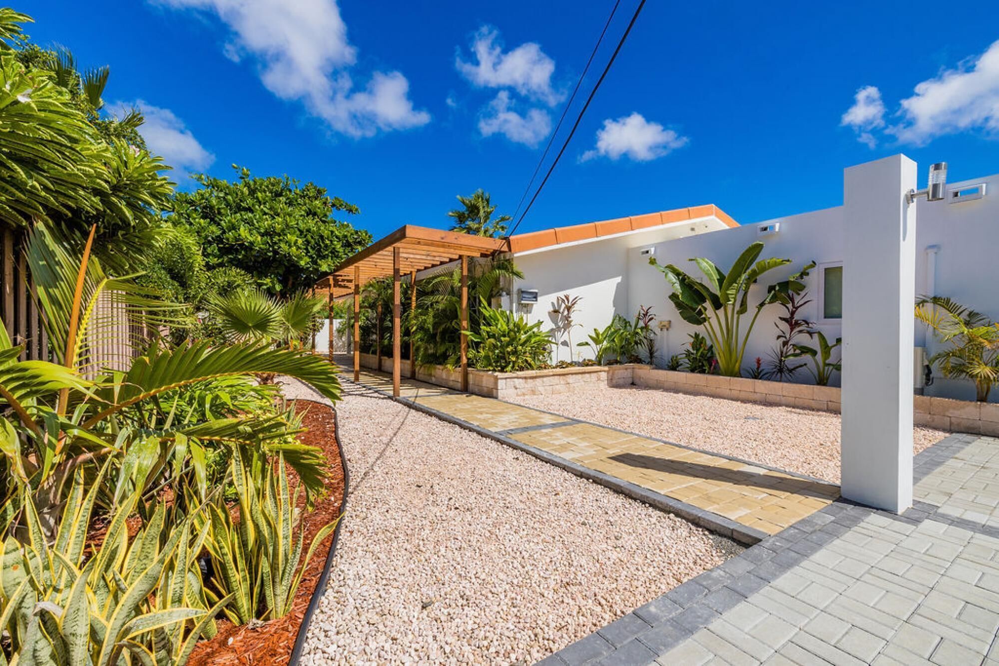 Um imóvel residencial com calçada pavimentada, pérgola de madeira e diversas plantas tropicais.