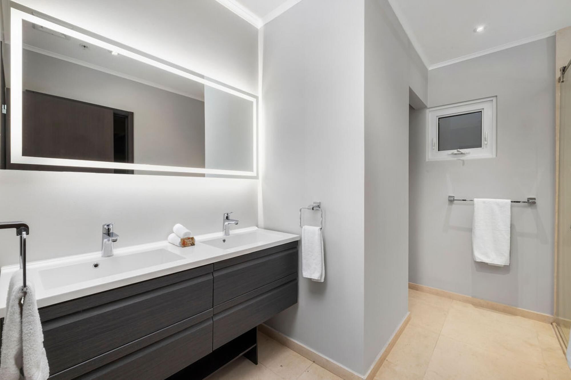 Un baño moderno con un espejo grande, dos lavamanos, muebles oscuros y una zona de ducha.