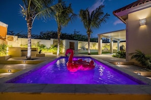 A swimming pool with a flamingo float, surrounded by palm trees and a covered patio area.