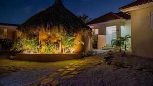 A thatched-roof structure with a well-lit garden, surrounded by a pathway and palm trees. - NEW Villa - Beautiful 3BR 2BA With Private Pool (Paradera)