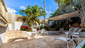A patio with stone flooring, white outdoor furniture, and a variety of plants and flowers. - 2BR 1BA Ground Level in the Heart of Aruba Pool (Noord)