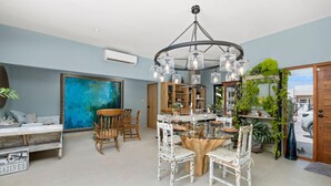 A spacious living room with a round wooden table, white chairs, a large abstract painting, and a ceiling light fixture. - 2BR Private Pool Outdoordining Amazing Gardens (Noord)