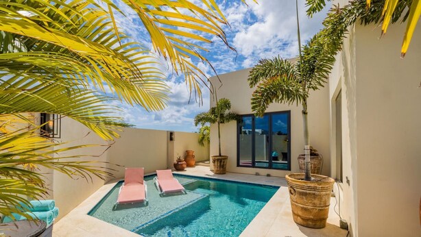 A pool area with two pink lounge chairs, a large planter, and a glass door leading to a covered patio. - 2BR Private Pool Outdoordining Amazing Gardens (Noord)