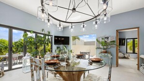 A modern dining area with a glass table, rustic chairs, and a chandelier. - 2BR Private Pool Outdoordining Amazing Gardens (Noord)
