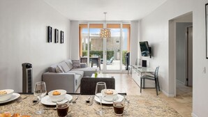 Een moderne woonkamer met een bank, eettafel en stoelen. Er is een televisie aan de muur bevestigd en een raam met uitzicht op het omliggende landschap.