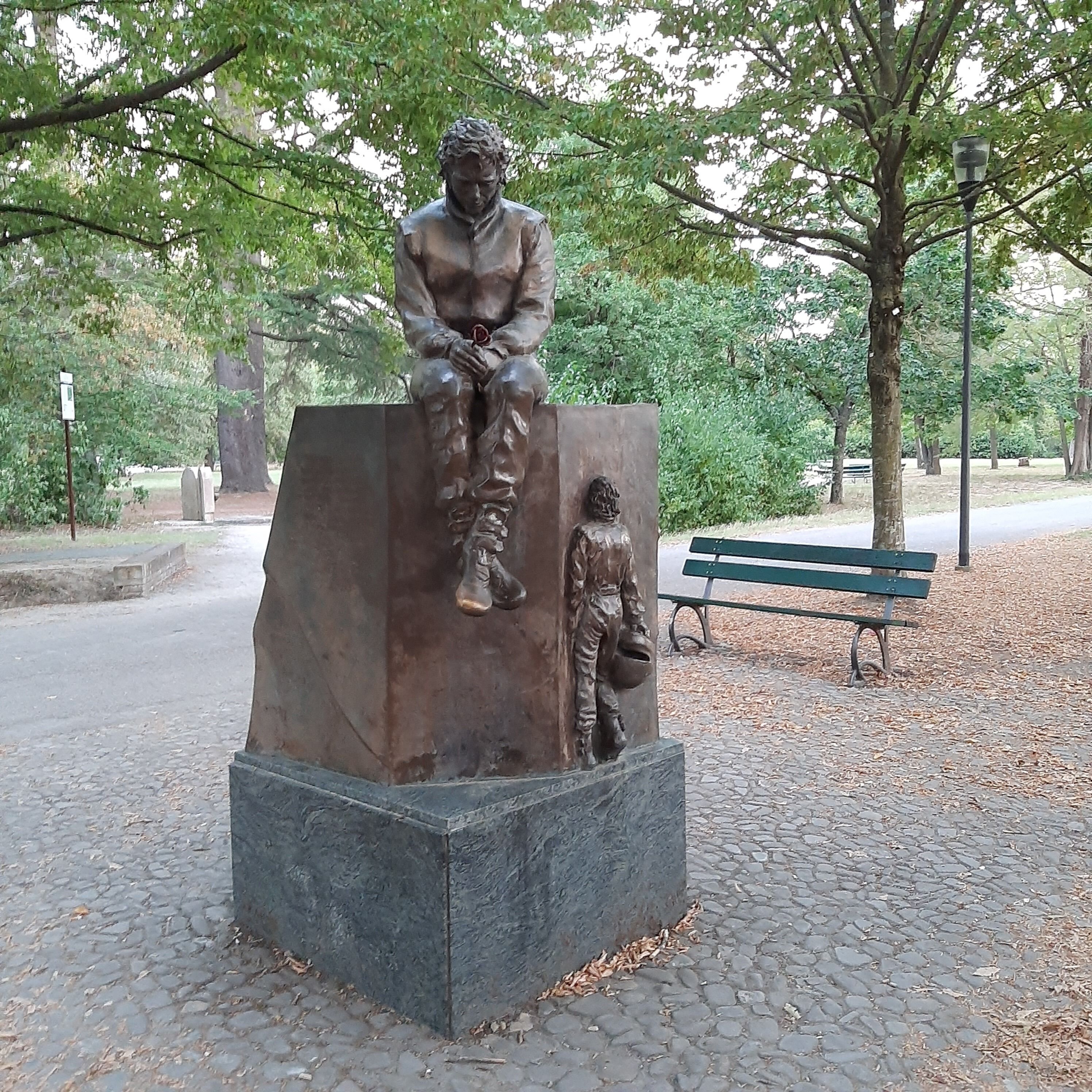 A statue of a seated figure on a pedestal in a park with a bench in the background.