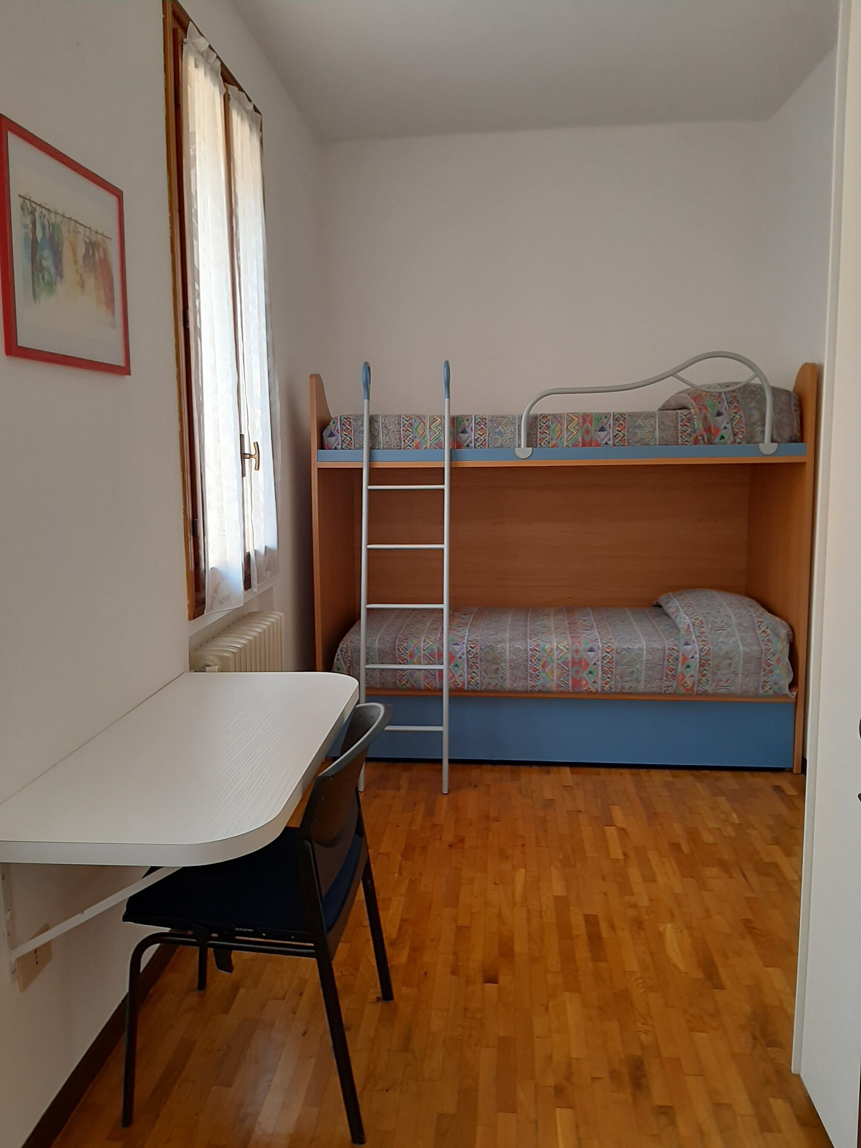 A small, well-lit room with a bunk bed, a desk with a chair, and a radiator.