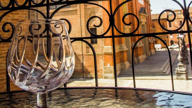 A clear glass vase on a table with a metal railing in the foreground, and a brick building with arched windows in the background.
