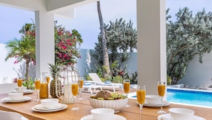 A dining table set with white plates, bowls, and glasses of orange juice, overlooking a swimming pool and lush greenery. - Brand NEW 3br2ba Villa With Pool in Great Location (Noord)