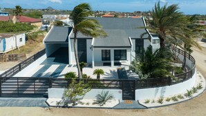 A modern house with a flat roof, large windows, and a fenced patio area with palm trees. - Brand NEW 3br2ba Villa With Pool in Great Location (Noord)
