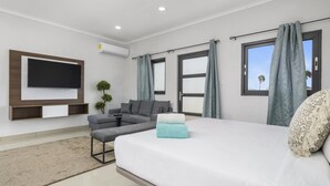 Ein modernes Hotelzimmer mit einem großen Bett, einem flachbildfernseher an der Wand, einer Sitzecke mit Sofa und Sesseln sowie großen Fenstern mit Vorhängen.