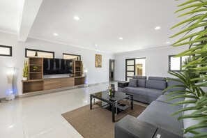 A modern living room with a grey sofa, a glass coffee table, and a wooden entertainment unit. - NEW 4br4ba Villa Across of Palmbeach w Pvt Pool (Noord)