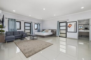 A modern hotel room with a sofa, a bed, a small table, and a bathroom with a mirror and sink. - NEW 4br4ba Villa Across of Palmbeach w Pvt Pool (Noord)