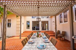 A dining area with a long table set for six, under a pergola with a lattice design.
