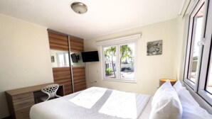 A bedroom with a bed, a desk, a chair, a TV, and a window with a view of palm trees. - Spacious 6BR Villa w Pool Close2beach Sleeps 16 (Noord)